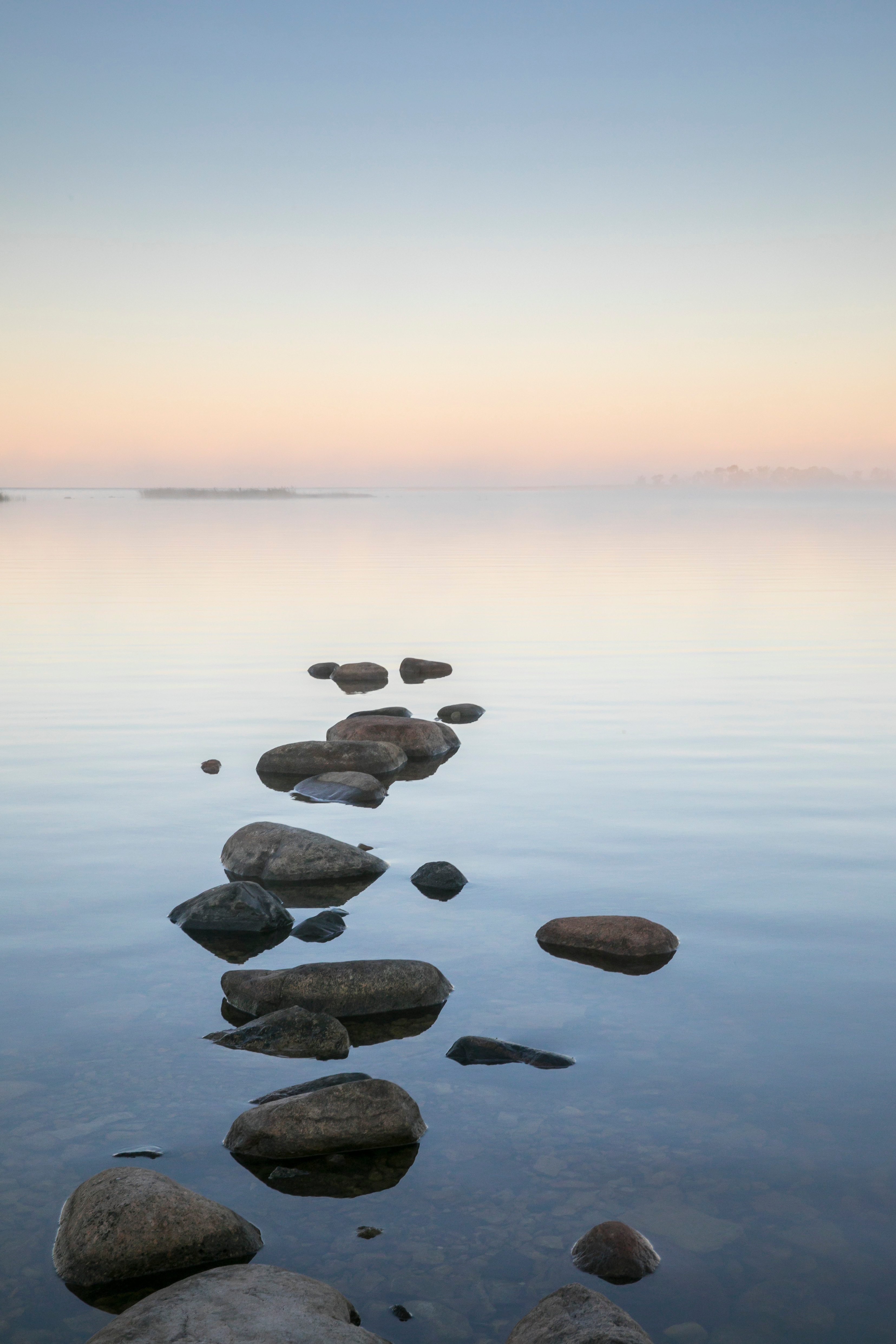 A calm scene of a trail of rocks peeking out of calm water with  an island and a peachy horizon in the backgroundl