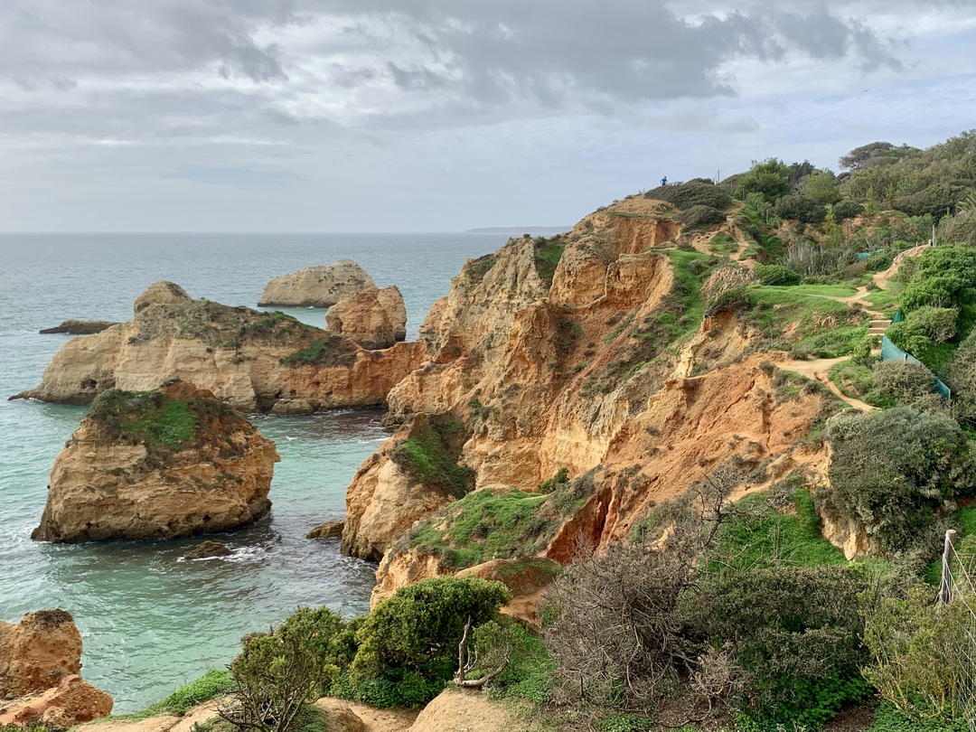 Sandy, orange colored bluffs with green vegetation overlooking blue water and grey sky.