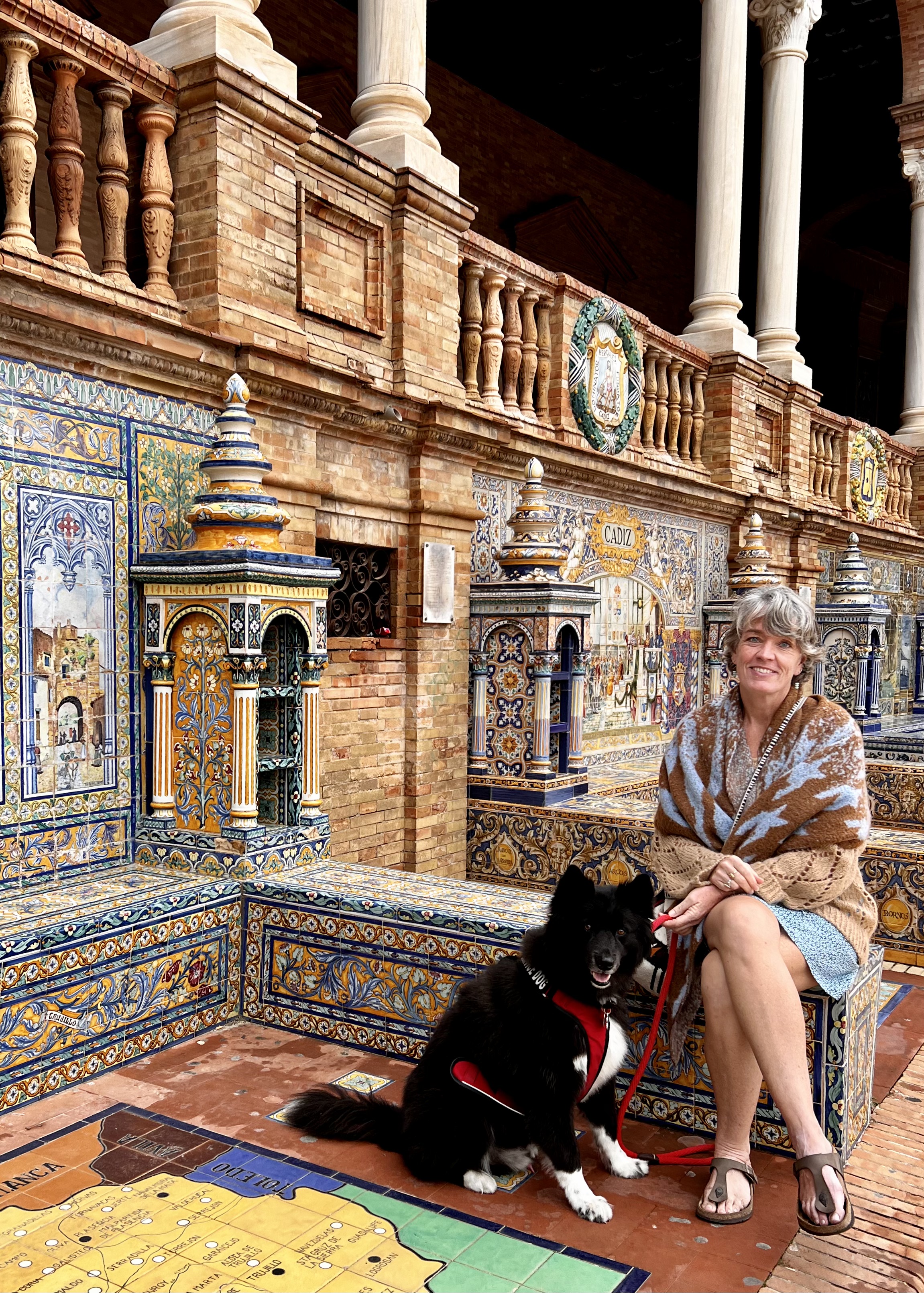 A woman and her dog sitting on a colorful tile bench next to a wall with a decorative tiled tower and colorful tiled scenes.